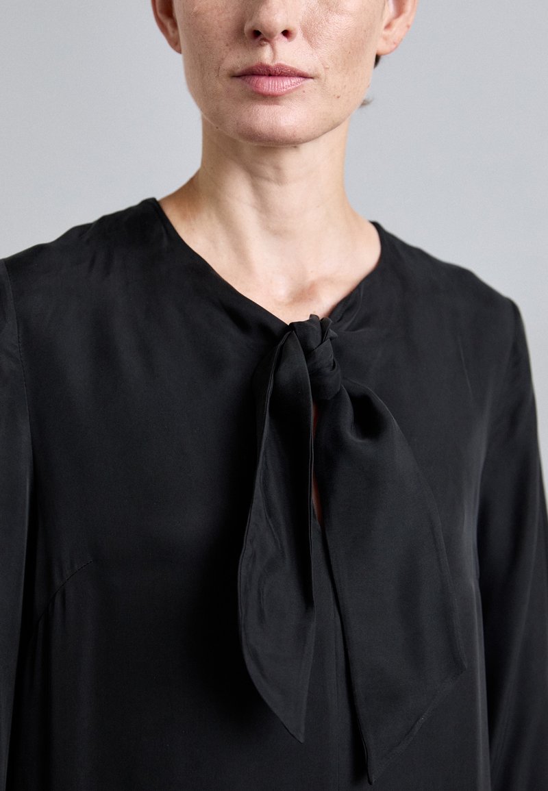 Schwarze Bluse mit rundem Ausschnitt, Bindung vorne und glatter Textur. Der Stoff wirkt leicht und hat einen weichen Fall.