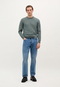 Un pull en laine gris, un jean bleu et des chaussures noires. Le modèle se tient contre un fond clair, les mains dans les poches. Coupe décontractée avec texture visible.