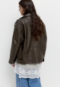 Frau mit langen braunen Haaren, die eine dunkelbraune Lederjacke über einem grauen Pullover und einem weißen Spitzenoberteil trägt, kombiniert mit blauen Jeans und seitlich steht.