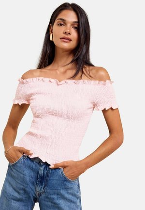 Frau trägt ein hellrosa schulterfreies, strukturiertes Oberteil und blaue Jeans mit den Händen in den Taschen, posiert vor einem schlichten Hintergrund.