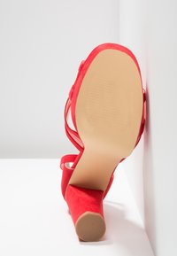 Sandale rouge à talon haut avec un talon bloc et une bride à la cheville, semelle tournée vers l'avant sur un fond blanc avec des angles.