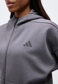 Femme portant un sweat à capuche gris zippé, avec un logo Adidas noir sur la poitrine gauche sur un fond uni.