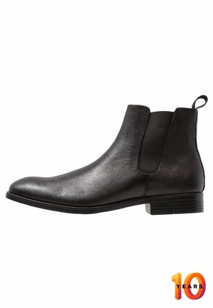 Botín Chelsea de cuero negro con acabado texturizado, paneles laterales elásticos y una lengüeta. Tacón bajo y diseño de punta redondeada.