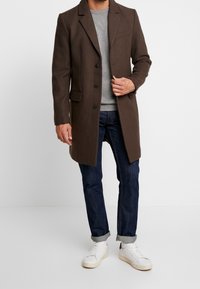 Brauner Wollmix-Mantel mit Revers, drei Knöpfen und seitlichen Taschen, getragen über einem grauen Pullover, kombiniert mit gerollten Jeans und weißen Turnschuhen.