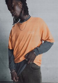 Orange kortærmet t-shirt lagdelt over en grå langærmet skjorte; inkluderer et lille logo og flere sølvtilbehør og ringe.