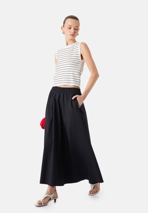 Nainen, jolla on valkoisessa, hihattomassa raidallisessa paidassa ja pitkä musta hame, seisoo toinen käsi taskussa ja valkoiset korolliset sandaalit jalassa.