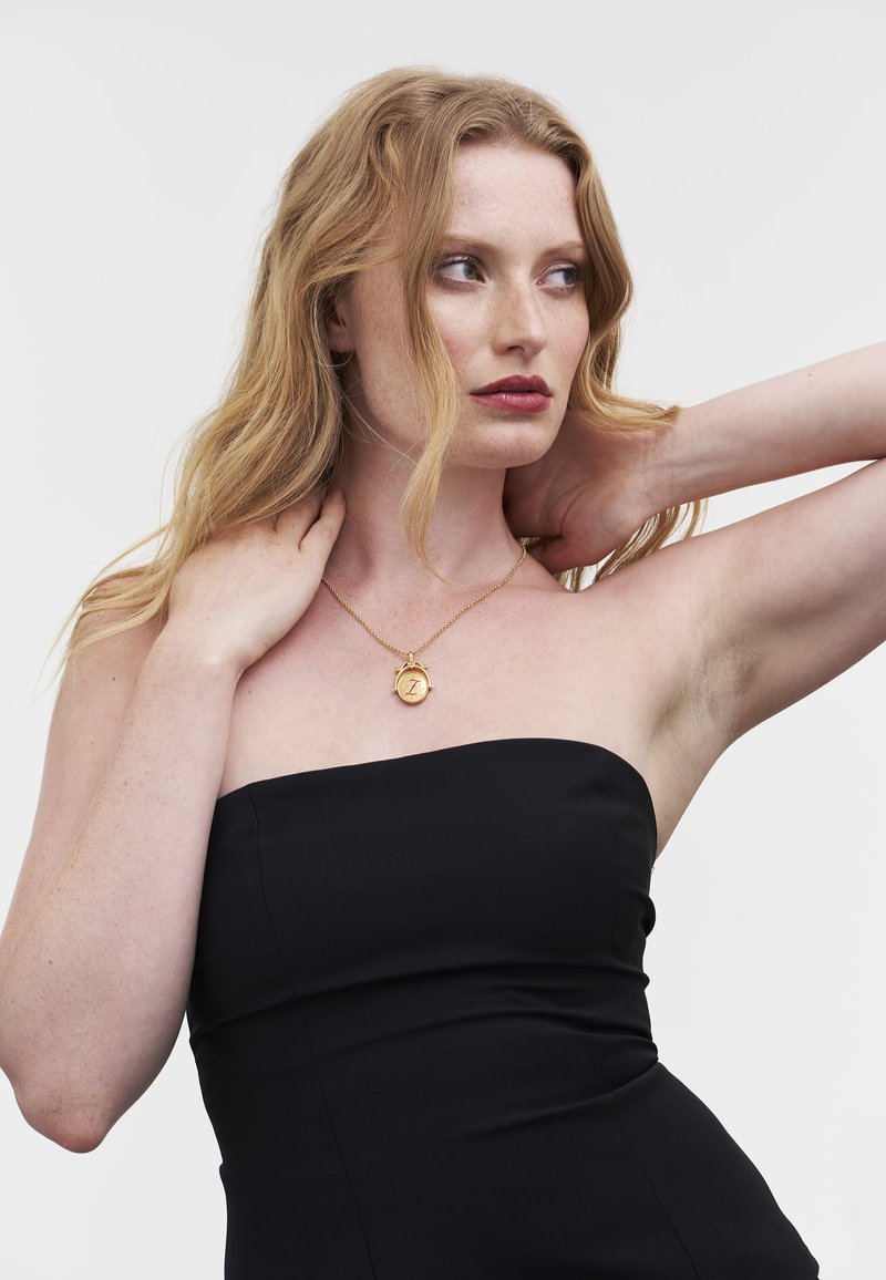 Gouden hangerketting aan een ketting, met een ronde hanger met een reliëfpatroon. Het model draagt een strapless zwarte jurk en heeft lang haar.