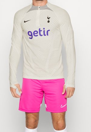 Athlète masculin portant un maillot à manches longues beige texturé du Tottenham Hotspur avec le logo violet "getir" et un short Nike rose vif.