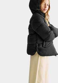 Giacca a vento nera con cappuccio, caratterizzata da un design trapuntato, bottoni visibili all'orlo e una texture di tessuto liscia. Abbinata a pantaloni beige.