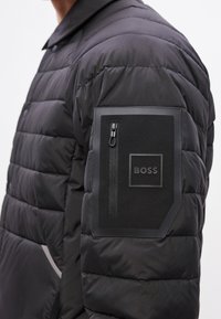 Fekete steppelt kabát, bal karján cipzáras zsebbel, texturált anyagból és dombornyomott logóval ellátott folttal.