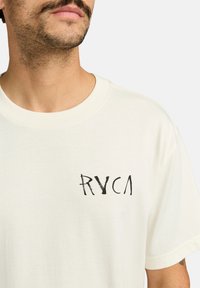Camiseta color crema con cuello redondo, con un logo gráfico negro "RYCA" en el área del pecho izquierdo. Tejido de algodón suave.