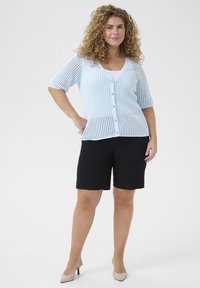 Cardigan en tricot bleu clair à manches courtes et boutons en perles, associé à un short noir et des talons hauts beiges. Texture lisse et design ajusté.