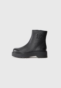 Bottine en cuir noir avec semelle épaisse et logo DKNY en relief sur le côté extérieur, présentée sur un fond uni.