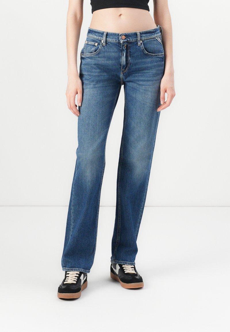 Jeans de mezclilla azul claro con un corte recto, diseño de cinco bolsillos y cierre de botón en la cintura. Llevados con zapatillas negras.