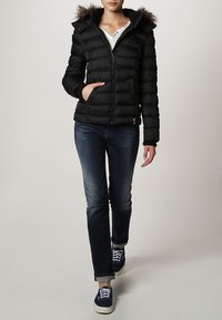 Black puffer jacket with a zip front, fur-lined hood, and diagonal quilted design. Paired with blue denim jeans and dark blue sneakers.