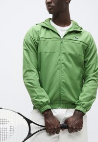 Zelena jakna z zadrgo iz lahkega blaga, z ruto in elastičnimi manšetami. Model drži teniško lopar, pod njo nosi krem hlače.