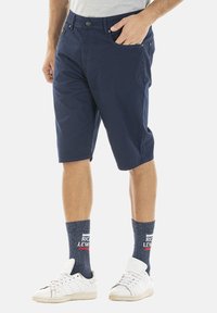 Bermuda en coton bleu marine avec une coupe droite, des poches latérales et une fermeture à bouton. Associé à des baskets blanches et des chaussettes à motifs bleus.