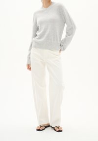 Personne portant un pull en V gris clair, un pantalon blanc à jambes larges et des tongs noires, debout avec une main dans la poche.