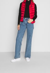 Chaleco de plumón rojo con cuello alto y cremallera, llevado sobre un suéter negro de cuello alto. Combinado con unos vaqueros de pierna ancha azules y zapatillas blancas.