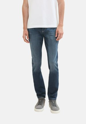 Jeans de denim azul con un ajuste slim, que presentan un lavado desvanecido, diseño de cinco bolsillos y costuras en contraste, usados con zapatillas grises.