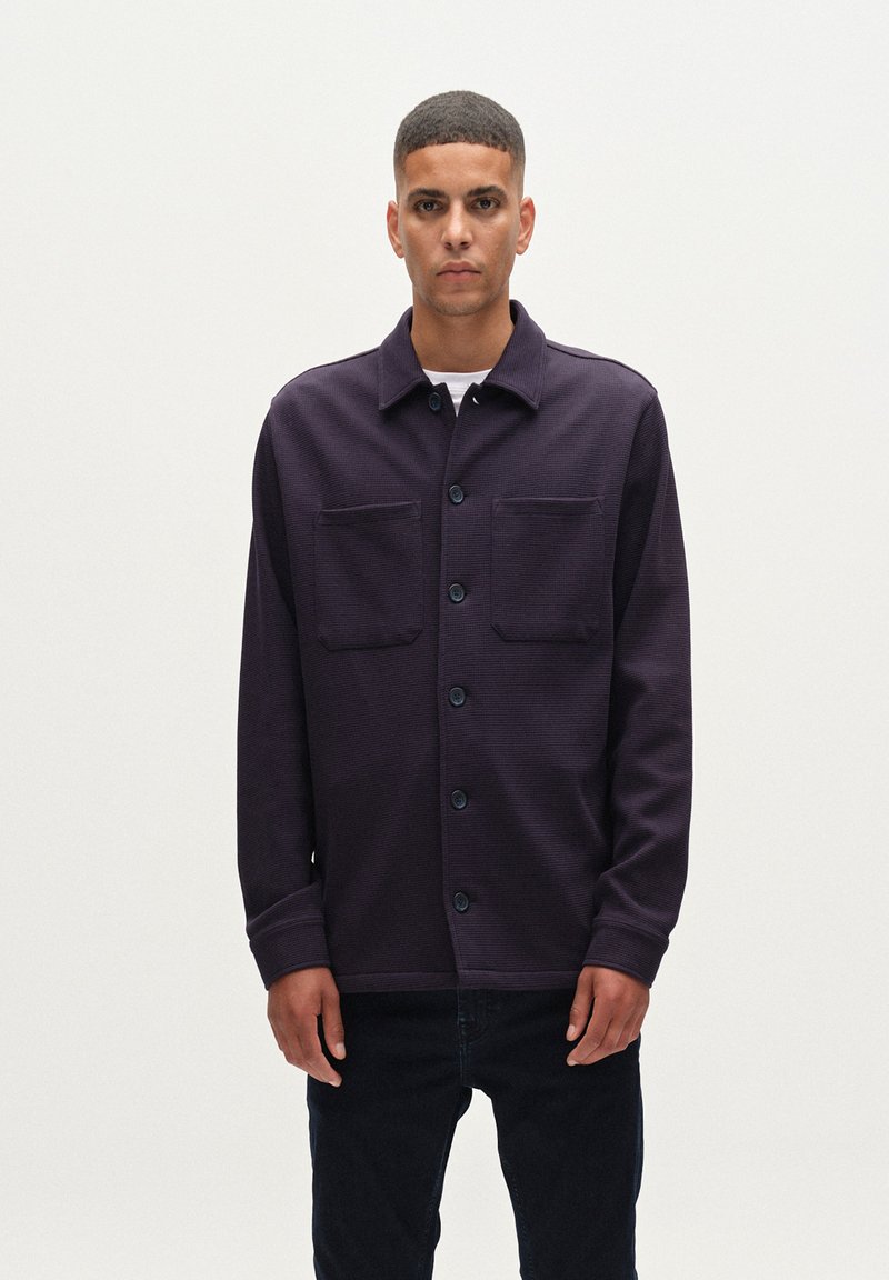 Camisa de botones azul marina hecha de tela texturizada, con dos bolsillos en el pecho y un cuello, combinada con jeans oscuros.