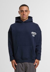 Tamsiai mėlyna medvilninė džemperis su priekiniais kišenėmis, plačia kapuce, trumpomis rankovėmis ir balta teksto logotipu ant krūtinės. Minkštas paviršius, kasdienis dizainas.