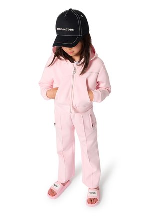 Pink zip-up hoodie with kangaroo pockets, matching pink pants with side zippers, and black cap, complemented by pink slide sandals.