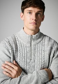 Suéter de cremallera gris claro con diseño de punto texturizado, cuello de canalé y patrones de trenza intrincados. Mangas largas con puños de canalé.