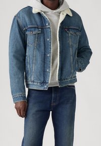 Uomo che indossa una giacca di jeans blu con collo in shearling sopra un hoodie grigio chiaro e jeans blu scuro, con una mano nella tasca della giacca.