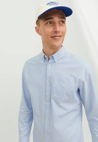 Hellblau-weiß gestreifte Button-up-Bluse mit Brusttasche, getragen mit einer cremefarbenen und blauen Kappe, auf der der Text "ORIGINALS CO. ATELIER" steht.