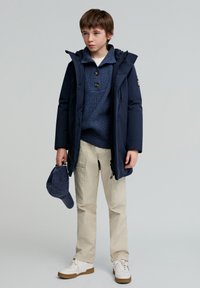 Marineparka med hætte, blå striktrøje, beige bukser og hvide sneakers. Holder en marinekasket. Glat tekstur, alsidig design, afslappet stil.