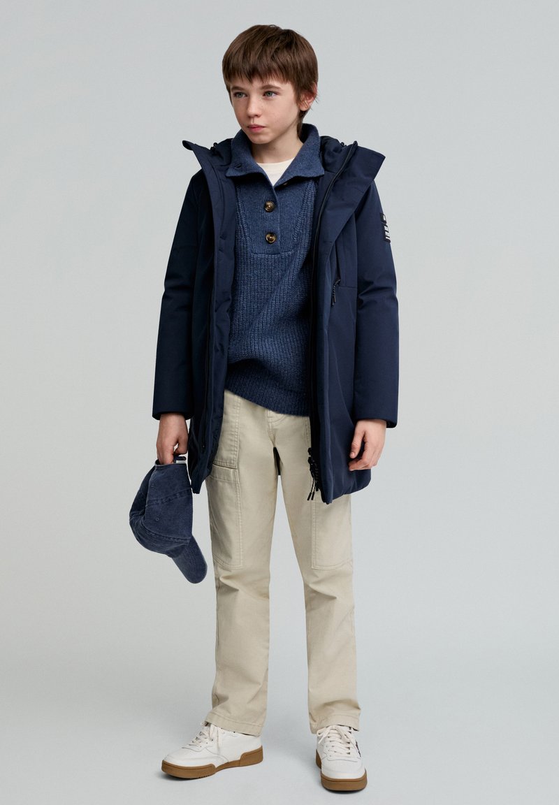 Marineparka med hætte, blå striktrøje, beige bukser og hvide sneakers. Holder en marinekasket. Glat tekstur, alsidig design, afslappet stil.