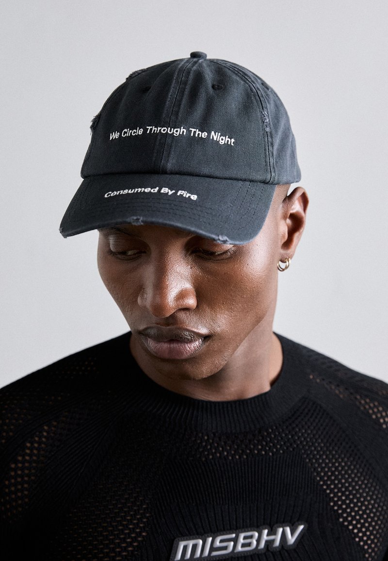 Casquette de baseball noire avec un texture en denim vieilli ; présente un texte blanc : "Nous tournons dans la nuit" et "Consommé par le feu."