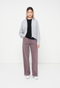 Cardigan grigio chiaro lavorato a maglia con motivo a trecce, sopra una camicia nera, abbinato a pantaloni larghi di un rosa polveroso e sneakers bianche.