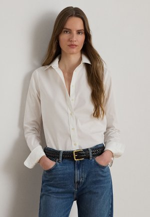 Une femme porte une chemise blanche boutonnée, un jean bleu et une ceinture noire tressée. Elle se tient debout, les mains dans les poches, devant un mur uni.