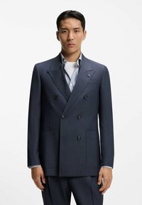 Giovane uomo che indossa una giacca doppiopetto blu scuro sopra una camicia azzurra e una maglietta bianca, in piedi contro uno sfondo bianco.