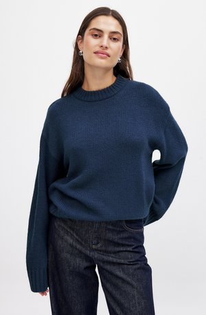 Suéter de punto azul oscuro con cuello redondo, hombros caídos y puños de canalé. Combinado con jeans de mezclilla oscuros, que muestran un corte relajado.