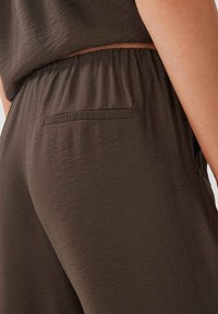 Vêtement texturé de couleur marron foncé doté d'une taille élastique et d'une petite poche plaquée à l'arrière. Design minimaliste avec une coupe décontractée.