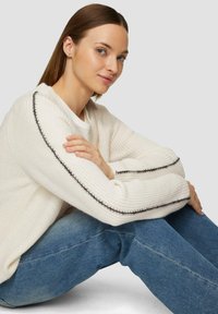 Gruby, kremowy sweter dziany o prążkowanej fakturze z kontrastującym czarnym przeszyciem wzdłuż rękawów, zestawiony z niebieskimi jeansami.