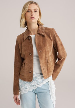 Femme portant une veste boutonnée en daim marron sur un haut blanc côtelé avec détail en dentelle et un jean bleu clair, debout devant un fond neutre.