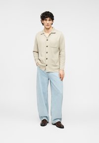 Φωτεινό μπεζ πουκάμισο με κουμπιά και μία τσέπη, συνδυασμένο με άνετο ανοιχτό μπλε τζιν και καφέ παπούτσια.