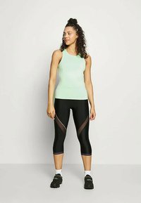 Top atlético sem mangas de verde claro, combinado com leggings pretas até ao meio da panturrilha, com painéis transparentes e detalhes curvados em castanho.