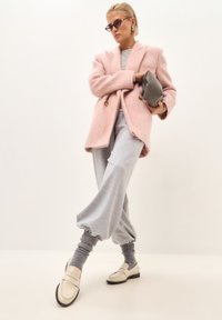 Rosa, flauschiger Mantel, graue, übergroße Sweatpants und cremefarbene Loafers mit schwarzen Sohlen. Modell hält eine graue Clutch und trägt runde Sonnenbrillen.