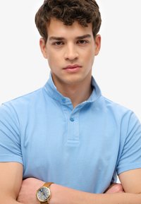 Lichtblauw poloshirt met korte mouwen en een opstaande kraag, voorzien van twee knopen en een gladde stoftextuur. Gouden horloge met een zwart wijzerplaat.