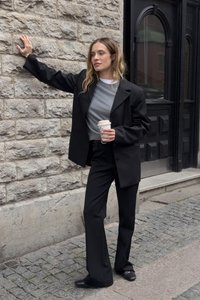 Ženska v črnem suknjiču in hlačah, sivo bluzo, drži skodelico kave in se opira ob kamnitem zidu na tlakovani ulici blizu črnih vrat.