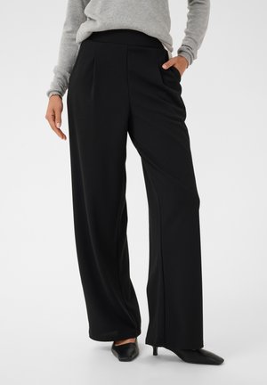 Femme portant un pantalon noir large, un pull gris et des chaussures noires à talons bas pointus, debout avec une main dans la poche.