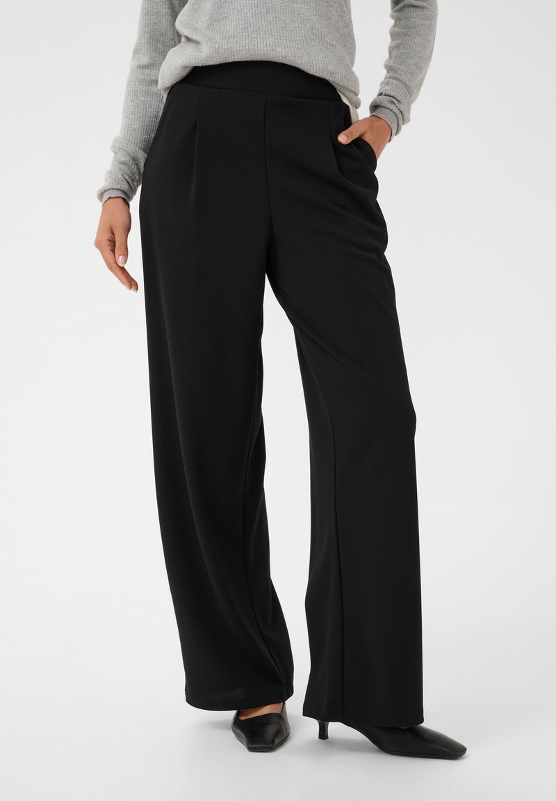 Femme portant un pantalon noir large, un pull gris et des chaussures noires à talons bas pointus, debout avec une main dans la poche.