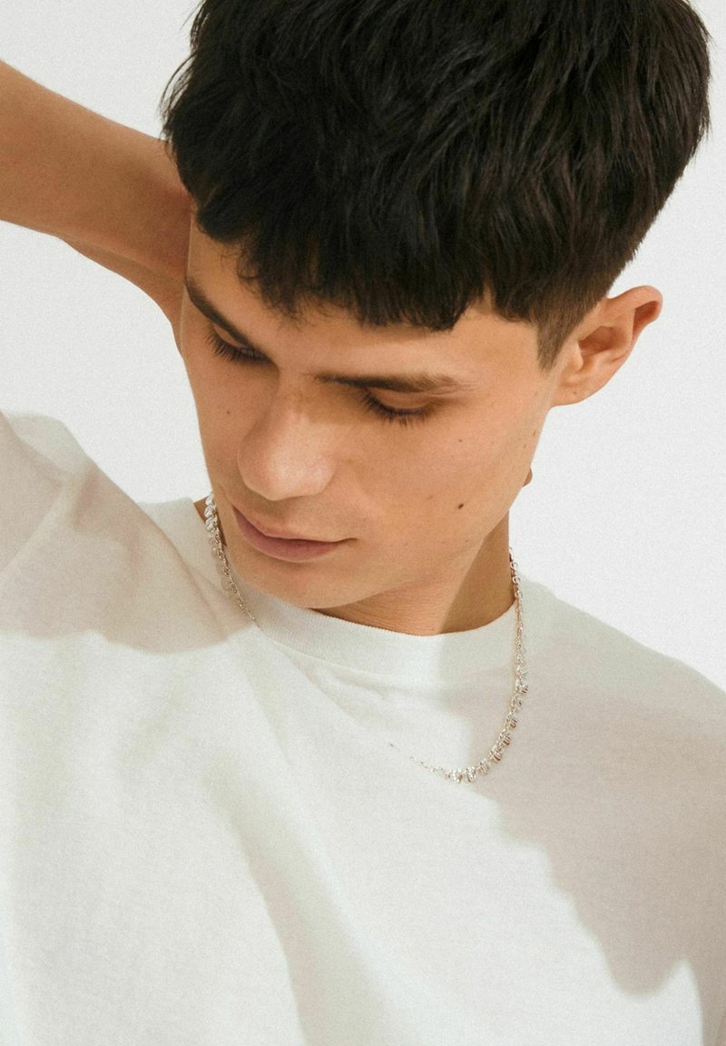 Joven con pelo corto y oscuro mirando hacia abajo, vistiendo una camiseta blanca y una delicada cadena de plata, con la mano detrás de la cabeza.