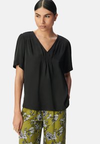 Schwarze Kurzarmbluse mit V-Ausschnitt, mit Raffung an den Schultern, kombiniert mit grünen Blumenmusterhosen.
