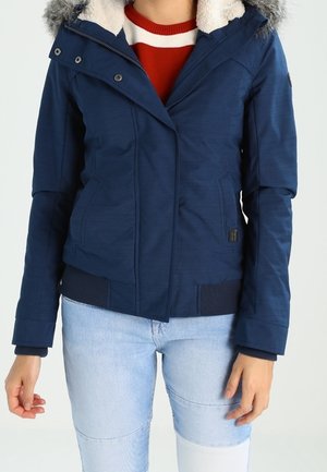 Persoon draagt een marineblauwe gevoerde winterjas met capuchon en bontkraag, gecombineerd met lichtblauwe spijkerbroek, staand voor een effen achtergrond.